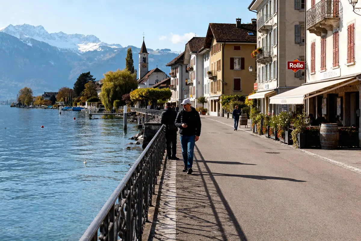 Promenade au bord d'un lac alpin, bâtiments historiques, montagnes enneigées en arrière-plan, personnes marchant.