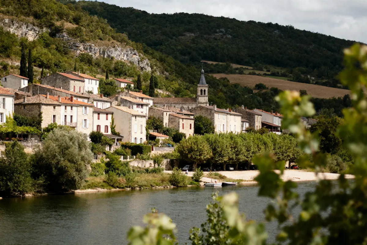 Village pittoresque au bord d'une rivière, maisons anciennes, arbres et collines verdoyantes en arrière-plan.